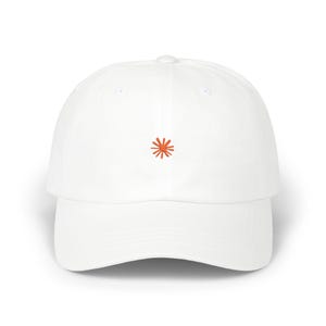 May include: A white baseball cap featuring a small, embroidered orange sunburst or star-shaped icon centered on the front panel. The cap has a structured crown, a curved brim, and a button at the top, maintaining a clean and simple aesthetic.