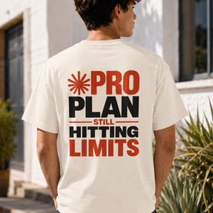May include: An off-white short-sleeve t-shirt featuring a graphic on the back with a red starburst icon and the text PRO PLAN STILL HITTING LIMITS in bold red and black lettering. The shirt has a crew neckline and a standard fit.