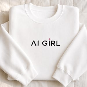 Op de afbeelding: Een wit sweatshirt met ronde hals en de geborduurde tekst AI GIRL in het zwart, met een klein roze vierpuntig sterretje boven de letter I. Het kledingstuk heeft lange mouwen, een geribbelde kraag en geribbelde boorden, in een gevouwen lay-out.