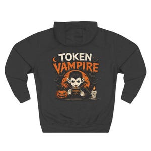 May include: A dark charcoal grey hooded sweatshirt featuring a graphic print of a vampire holding a mug labelled "TOKENS </>". The design includes an orange moon, a jack-o'-lantern, a melting candle, and bats, with the text "TOKEN VAMPIRE" above.