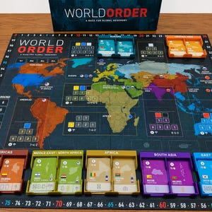 Puede incluir: Un juego de mesa World Order que incluye un tablero con un mapa mundial de fondo oscuro con bordes numerados, bandejas de cartas regionales de colores y la tapa de la caja con el título World Order: A Race for Global Hegemony en blanco y rojo.