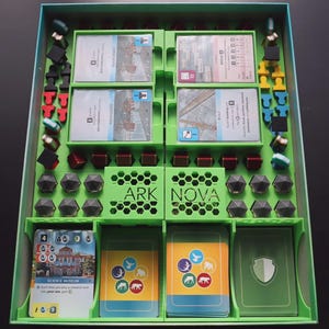 Op de afbeelding: Een bordspel-organizer inzetstuk van felgroen plastic, met gelabelde vakken voor Ark Nova spelonderdelen. Het bevat sleuven voor kaartspellen, kleine rode en zwarte blokjes, grijze zeshoekige fiches en diverse gekleurde speelfiguren.