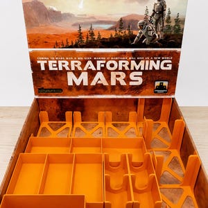 May include: The Terraforming Mars board game box features an open lid displaying a Martian landscape with two astronauts and the text "Coming to Mars was a big step. Making it habitable will give us a new world." The interior contains orange plastic inserts.