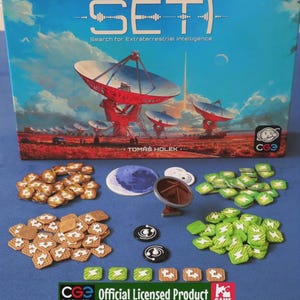 Puede incluir: El juego de mesa SETI: Search for Extraterrestrial Intelligence muestra una caja con antenas parabólicas rojas en un paisaje. Los componentes incluyen fichas cuadradas marrones y verdes, dos fichas negras circulares, un disco lunar y una antena miniatura.