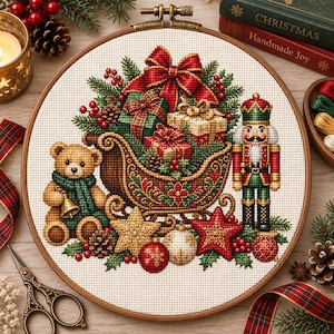 Op de afbeelding: Een ronde borduurring met een kruissteekontwerp van een bruine teddybeer met een groene sjaal, een houten slee gevuld met ingepakte cadeaus en een notenkraker-soldaat. De scène bevat rode en gouden ornamenten en sterren.