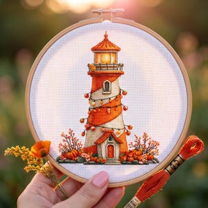 Puede incluir: Bordado de punto de cruz de un faro con rayas naranjas y blancas, un tejado rojo y una pequeña casa en la base. El faro está decorado con luces de cadena y rodeado de calabazas y follaje otoñal, en un bastidor de bordado de madera.
