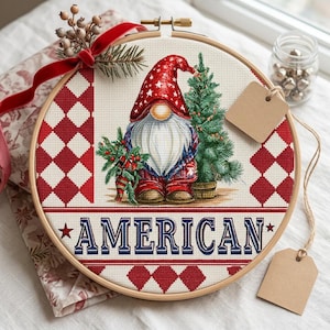 May include: A cross-stitch embroidery hoop featuring a gnome wearing a red and white star-patterned hat and a patriotic outfit. The word "AMERICAN" is displayed in blue text. The design includes a small Christmas tree and red and white diamond patterns.