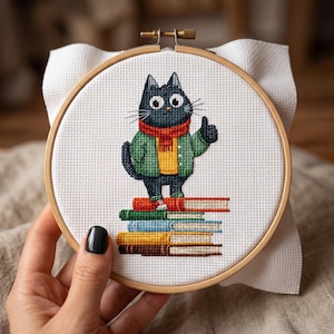 May include: Cross-stitch embroidery in a wooden hoop features a black cat wearing a green jacket and red scarf, giving a thumbs-up. The cat stands on a stack of books with various colored spines. The background is white.
