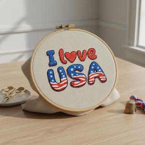 May include: A cross-stitch embroidery hoop with the text "I love USA" in red, white, and blue. The embroidery is in a wooden hoop, with a beige fabric background. Gold scissors, a thimble, and embroidery floss are also visible.