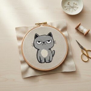 May include: A cross-stitch embroidery hoop featuring a grumpy gray cat with a white belly spot. The cat is stitched on a beige fabric, set in a wooden hoop. Additional craft supplies, including pins, thread, and scissors, are visible.