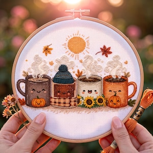 May include: A circular embroidery hoop containing a cross-stitch design of four smiling mugs with steam, a yellow sun, and autumn leaves. The mugs feature a pumpkin, a blue knit hat with a scarf, sunflowers, and a small pumpkin on a white fabric.