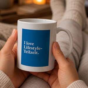 Könnte beinhalten: Weißer Keramikbecher mit blauem Quadrat und dem Text "I love Lifestyle-Teilzeit." Der Becher hat einen gebogenen Henkel und wird mit beiden Händen gehalten. Der Hintergrund ist unscharf, was auf eine gemütliche Umgebung hindeutet.