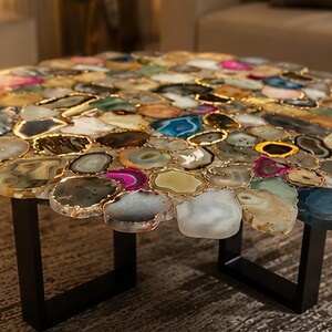 May include: A round coffee table with a mosaic top made of colorful agate slices. Each slice is outlined in gold and set on a black metal base. The table's design features a variety of colors, including blue, pink, and green.