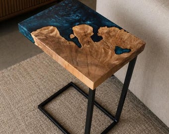 Op de afbeelding: Een rechthoekige bijzettafel met een natuurlijk houten blad met een levendige rand en een opvallende nerf, gecombineerd met een diepblauw doorschijnend harsgedeelte. Het blad rust op een zwart metalen C-vormig frame.