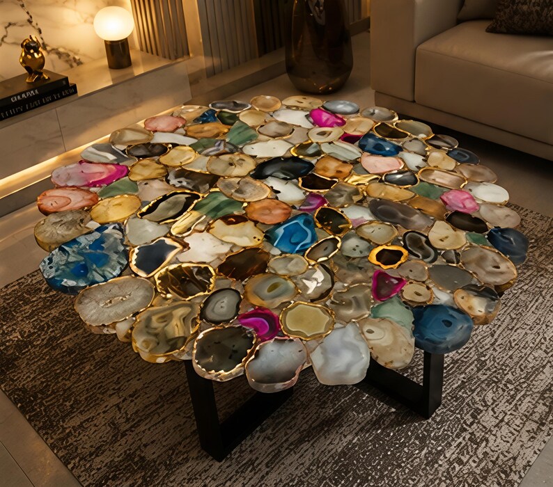 May include: A round coffee table with a mosaic top made of various colorful agate slices. Each slice is outlined in gold and set on a black metal base. The table's design incorporates shades of blue, pink, and yellow.