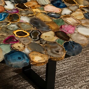 May include: A round table with a mosaic top made of polished, irregularly shaped stones in shades of blue, pink, brown, and gold. The stones are set in a gold-colored border. The table has a black metal base.