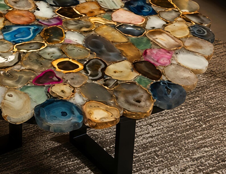 May include: A round table with a mosaic top made of polished, irregularly shaped stones in shades of blue, pink, brown, and gold. The stones are set in a gold-colored border. The table has a black metal base.