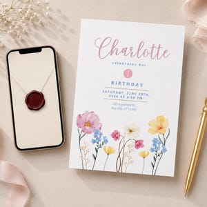 Può includere: Un invito di compleanno bianco con fiori di campo ad acquerello sul fondo e testo rosa Charlotte celebrates her 1st birthday, Saturday, June 20th, 2026 at 4:00 PM, 123 Anywhere St., Any City, ST 12345, accanto a uno smartphone e una penna dorata.