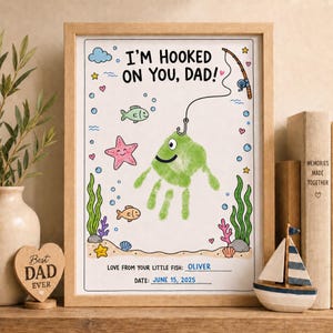 May include: A framed art print featuring the text I'M HOOKED ON YOU, DAD! with a green handprint transformed into a fish on a fishing line. The design includes colorful illustrations of a starfish, small fish, seaweed, and bubbles, with space for a name and date.