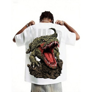 May include: White t-shirt featuring a detailed graphic of a green and brown alligator with a red open mouth. The alligator is on a brown rock. The word "WILD" is printed in white on the back of the shirt.