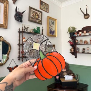 Peut inclure: Un attrape-soleil en vitrail en forme de citrouille avec des panneaux de verre orange vif, une petite tige verte et un fond en verre clair texturé avec un accent central en forme de losange, le tout assemblé par des lignes de soudure en métal sombre.