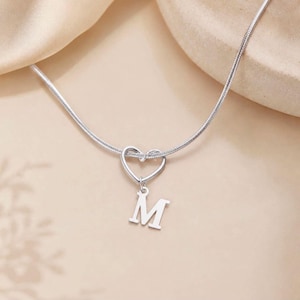 Peut inclure: Un collier argenté avec un pendentif en forme de cœur et la lettre "M" suspendue. La chaîne est fine et le pendentif est de conception simple. Le fond est de couleur neutre et douce. Un bijou délicat.