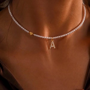 Peut inclure: Un collier de perles délicat avec un charm en forme de cœur doré et un pendentif doré en forme de lettre "A", incrusté de petites pierres scintillantes. Le collier est porté autour du cou.