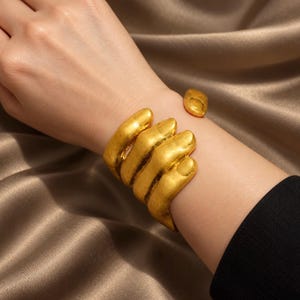 Peut inclure: Un bracelet manchette doré en forme de main humaine qui s'enroule autour du poignet. Le métal présente une finition mate et texturée avec des doigts et un pouce bien définis formant le design ouvert du bijou.