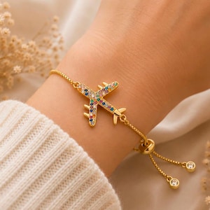 Peut inclure: Bracelet doré orné d'un charm en forme d'avion incrusté de pierres multicolores. Le bracelet est porté sur un poignet, avec une manche de pull blanche en arrière-plan. Le bracelet est réglable.