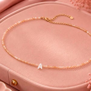 Könnte beinhalten: Eine zarte Halskette mit kleinen, hellorangen Perlen und einer goldfarbenen Kette. Die Halskette hat einen weißen Anhänger mit dem Buchstaben "A". Der Schmuck wird auf einer rosa Oberfläche mit rosa Stoff und floralen Akzenten präsentiert.
