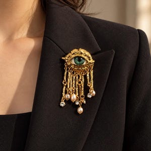 Peut inclure: Une broche dorée représentant un motif d'œil central avec un iris vert. L'œil est entouré d'un cadre texturé aux formes organiques. Des chaînes dorées ornées de petites perles blanches et de cristaux transparents pendent de la base.