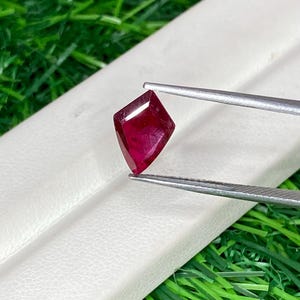 Könnte beinhalten: Ein drachenförmiger, tiefroter Edelstein, der von einer silberfarbenen Metallpinzette gehalten wird. Der Stein hat eine facettierte Oberfläche, die das Licht reflektiert und einen durchscheinenden, gesättigten karmesinroten Farbton zeigt.