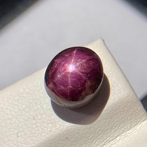 May include: A polished, oval-shaped gemstone with a deep burgundy color. The gem has a star-like effect, with lines radiating from a central point. The stone is set against a white surface, with a shadow cast to the right.