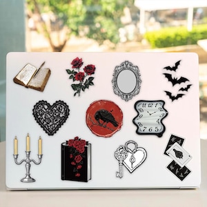 May include: A white laptop lid decorated with various stickers including an open book, red roses, an ornate mirror, black bats, a black lace heart, a red wax seal with a raven, a melting clock, a silver candelabra, a book with roses, a key, and cards.