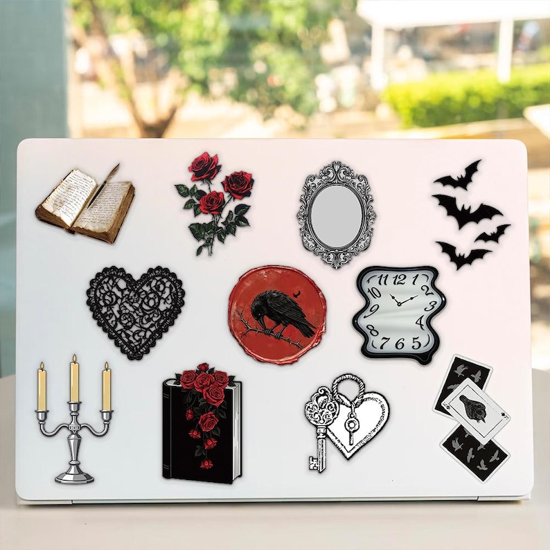 May include: A white laptop lid decorated with various stickers including an open book, red roses, an ornate mirror, black bats, a black lace heart, a red wax seal with a raven, a melting clock, a silver candelabra, a book with roses, a key, and cards.