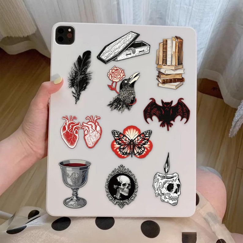 May include: A white tablet case decorated with various gothic-themed stickers including a black feather, an open coffin, a stack of books, a raven with a red rose, a bat, a pair of anatomical hearts, a butterfly, a silver goblet, a skull cameo, and a candle.