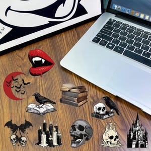 May include: A collection of gothic-themed stickers featuring red vampire lips, a red crescent moon with bats, a raven on an open book, a stack of old books, a raven on a skull, bat-winged skulls, black candles, a human skull, and a castle.