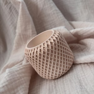 May include: A small, peach-colored planter with a geometric diamond pattern. The cylindrical pot has a textured surface and is set against a soft, neutral-toned fabric background. Ideal for small plants or succulents.