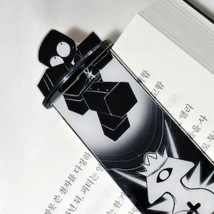May include: Black and white bookmark with a unique design featuring geometric shapes and cartoon-like characters. The bookmark is inserted into an open book with Korean text.