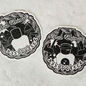 May include: Two circular stickers with a black and white glitter finish. Each sticker features a central blocky character, the text "Bad Things" at the top, the text "[4001]" at the bottom, and decorative heart icons along the lower edge.