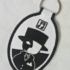 May include: An oval-shaped embroidered keyring featuring a black and white design of a figure wearing a top hat and a mask. Above the figure is a square icon containing a musical note symbol. The keyring has a black stitched border and a metal ring.