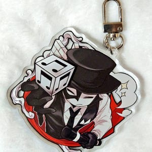 以下が含まれることがあります： 黒いシルクハット、白いマスク、白いシャツの上に黒いベストを着たキャラクターのアクリルキーホルダー。キャラクターは黒い音符が描かれた白いキューブとマイクを持っており、背景には赤い円がデザインされています。