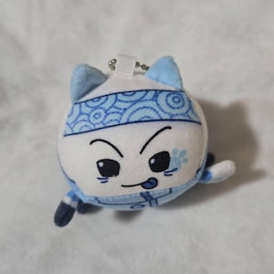 Puede incluir: Un llavero de peluche redondo con forma de gato, de pelaje blanco y azul claro. El personaje lleva una cinta azul con un patrón de ondas circulares, ojos negros bordados, una pequeña lengua fuera y una huella de pata azul en la mejilla.