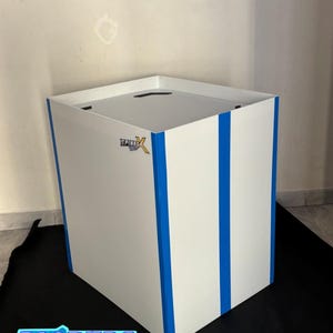 Può includere: Una scatola espositiva quadrata bianca con strisce blu verticali sui pannelli frontali e laterali. La superficie superiore è piatta e bianca con una fessura nera. Un piccolo logo con il testo SPINBOX è stampato sull'angolo anteriore superiore.
