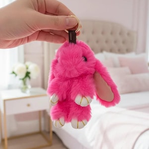 May include: A bright hot pink plush rabbit keychain featuring soft faux fur, long floppy ears with smooth pink inner linings, and small white paws with gold-colored stitched details. The rabbit is attached to a gold-toned metal ring.