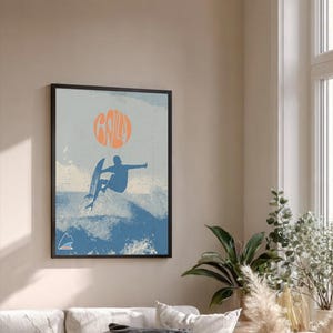 以下が含まれることがあります： 青と白の波を背景に、サーファーのシルエットが描かれた額入りのアートプリント。サーファーの上にはオレンジ色の「Gaily」の文字。レトロなスタイルで、海岸沿いのインテリアに最適です。