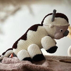 May include: A handmade crochet plush toy shaped like a bison with a cream-colored body, dark brown patches on the back and head, and dark brown feet. The creature features small horns, a rounded snout, and a long tail with a dark brown tip.