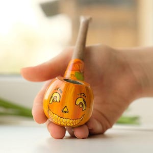 May include: A hand-painted, orange pumpkin-shaped pipe with a smiling face design. The pipe has a wooden stem and is held in a person's hand. The background is blurred, with a window and flowers visible.