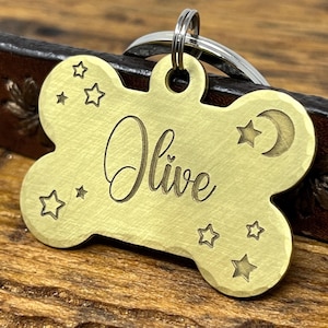 May include: Brass bone-shaped dog tag with the name "Olive" engraved in cursive script. The tag features a crescent moon and stars.