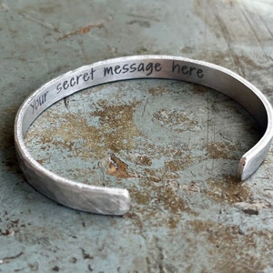 May include: A silver metal bracelet with a hammered finish. The bracelet is engraved with the text "Your secret message here".